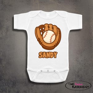 May include: White baby bodysuit featuring a baseball glove and ball graphic. The glove is brown, and the ball is white with red stitching. The name "SANDY" is printed in brown below the graphic. The Pink Basement logo is in the bottom right corner.