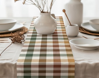 May include: A checkered table runner in shades of green, brown, and cream. The runner is placed on a white tablecloth, with white ceramic vases and plates in the background. Dried flowers and wooden utensils add to the rustic aesthetic.