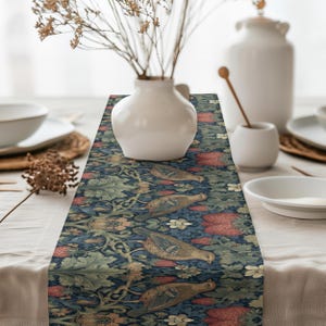 May include: A decorative table runner with a detailed floral and bird pattern. The runner features a dark blue background with green leaves, red flowers, and brown birds. It is placed on a white tablecloth, with white vases and tableware in the background.