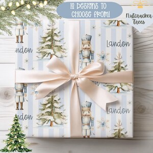May include: Wrapping paper featuring a light blue and white striped pattern with watercolor illustrations of nutcrackers, snow-covered pine trees, and snowflakes. The name Landon is printed in dark text. A pale pink ribbon is tied around the box.
