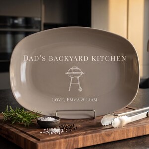 May include: An oval taupe serving platter featuring the white text DAD'S BACKYARD KITCHEN, a line drawing of a charcoal grill, and the text LOVE, EMMA & LIAM. The platter is displayed on a metal stand, accompanied by a small bowl of salt and tongs.