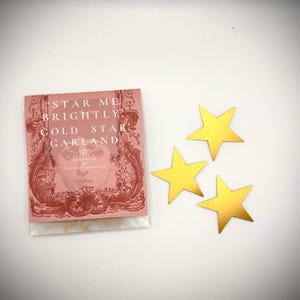 Może przedstawiać: Mały r&oacute;żowy pakunek z tekstem "Star Me Brightly Gold Star Garland" i słowami "Handmade by The Box and Alice" na nim. Trzy złote ozdoby w kształcie gwiazd znajdują się na białej powierzchni.