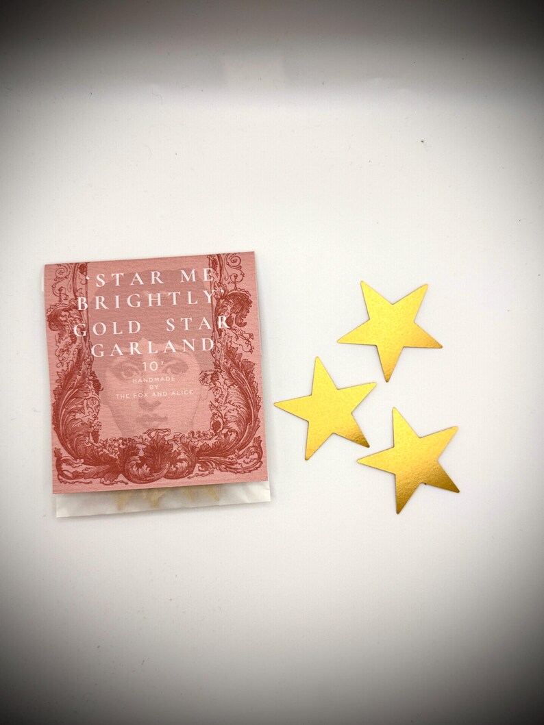 Może przedstawiać: Mały r&oacute;żowy pakunek z tekstem "Star Me Brightly Gold Star Garland" i słowami "Handmade by The Box and Alice" na nim. Trzy złote ozdoby w kształcie gwiazd znajdują się na białej powierzchni.