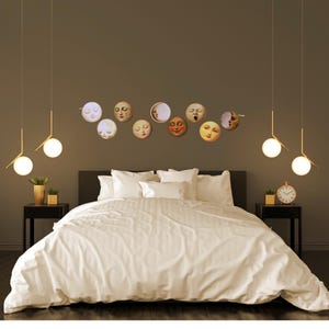 K&ouml;nnte beinhalten: Ein Schlafzimmer mit einem Bett mit wei&szlig;er Bettw&auml;sche, flankiert von schwarzen Nachttischen. &Uuml;ber dem Bett befindet sich eine Sammlung von Wandkunst mit Mondgesichtern. Goldene Pendelleuchten h&auml;ngen von der Decke, und eine kleine Uhr steht auf einem Nachttisch.