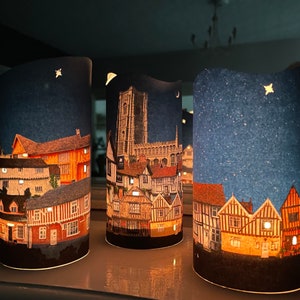 May include: Three cylindrical candle holders with a night sky background and a cityscape design. The cityscape features buildings with a mix of brick and timber framing. The candles are lit and glow through the paper.