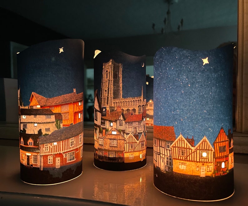 May include: Three cylindrical candle holders with a night sky background and a cityscape design. The cityscape features buildings with a mix of brick and timber framing. The candles are lit and glow through the paper.