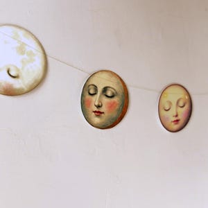 K&ouml;nnte beinhalten: Eine Girlande mit Mondphasen-Ausschnitten mit geschlossenen Augen und rosigen Wangen. Die Dekoration mit Himmelsmotiven zeigt einen Vollmond, einen Halbmond und Zwischenphasen, die auf einer d&uuml;nnen wei&szlig;en Schnur aufgereiht sind. Die Monde sind in weichen, ged&auml;mpften Farben gemalt.