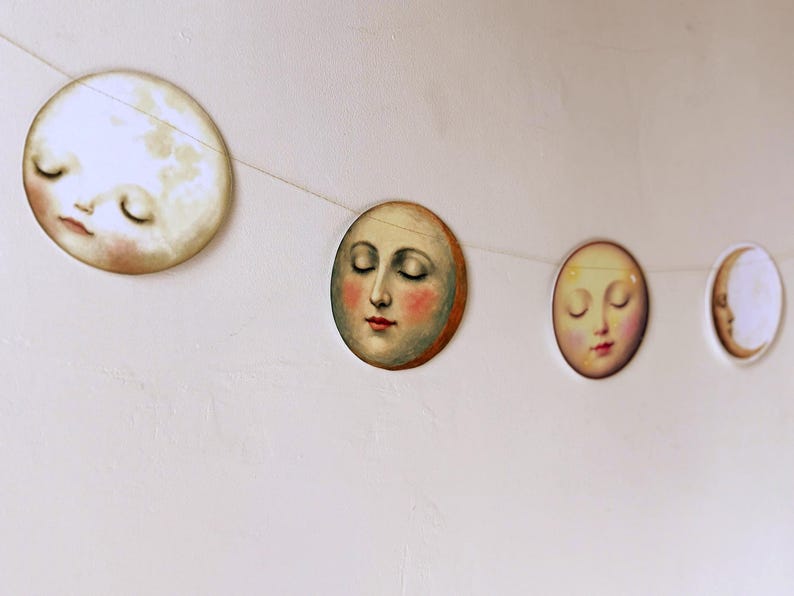 K&ouml;nnte beinhalten: Eine Girlande mit Mondphasen-Ausschnitten mit geschlossenen Augen und rosigen Wangen. Die Dekoration mit Himmelsmotiven zeigt einen Vollmond, einen Halbmond und Zwischenphasen, die auf einer d&uuml;nnen wei&szlig;en Schnur aufgereiht sind. Die Monde sind in weichen, ged&auml;mpften Farben gemalt.
