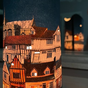 May include: A cylindrical candle holder with a printed design of a town at night. The design features a crescent moon, stars, and several buildings with lit windows. The candle holder is made of paper and has a white base.