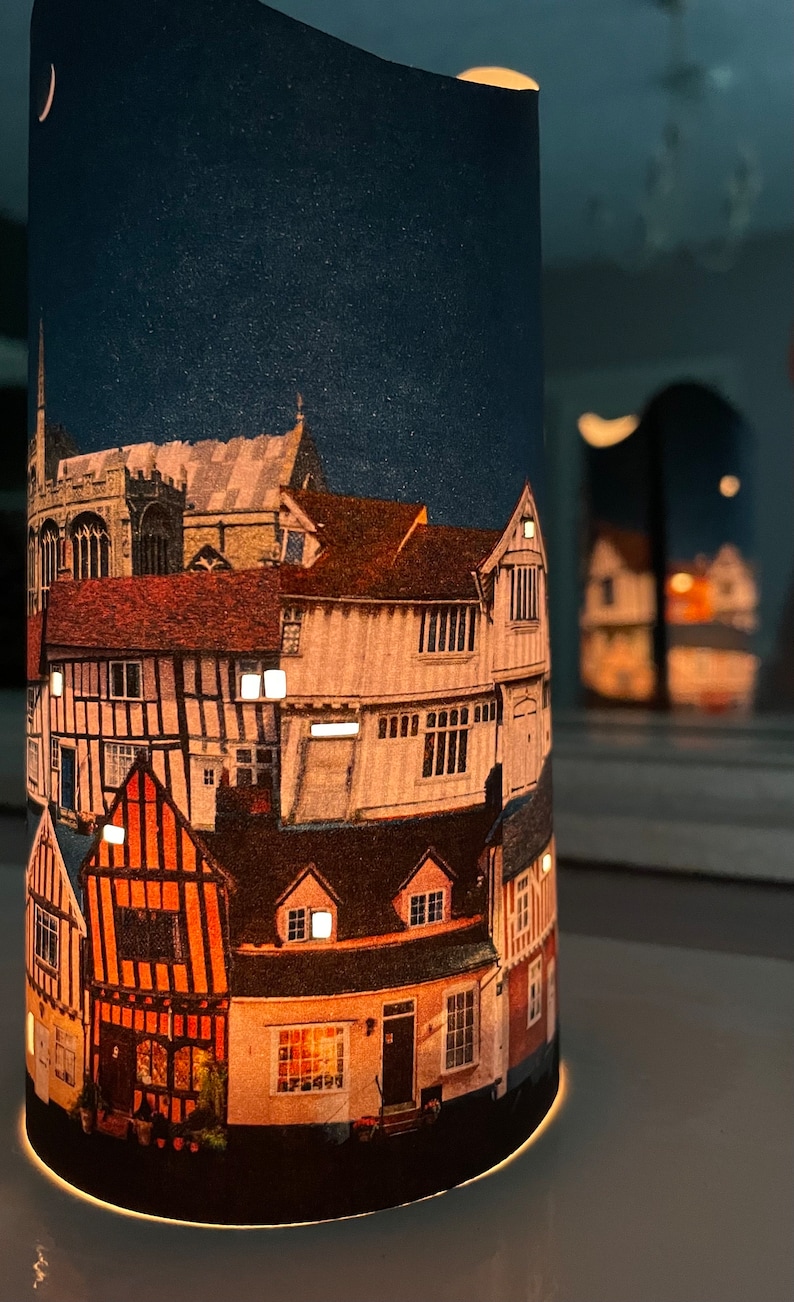 May include: A cylindrical candle holder with a printed design of a town at night. The design features a crescent moon, stars, and several buildings with lit windows. The candle holder is made of paper and has a white base.