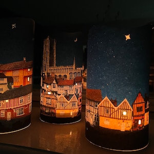 May include: Three cylindrical candle holders with a night scene printed on them. The first candle holder has a cityscape with a church in the background. The second candle holder has a cityscape with a crescent moon in the sky. The third candle holder has a cityscape with a star in the sky.