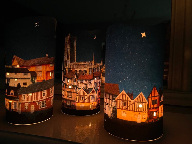 May include: Three cylindrical candle holders with a night scene printed on them. The first candle holder has a cityscape with a church in the background. The second candle holder has a cityscape with a crescent moon in the sky. The third candle holder has a cityscape with a star in the sky.