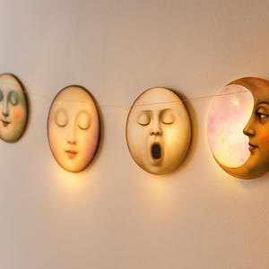 K&ouml;nnte beinhalten: Eine Lichterkette mit dekorativen Mondgesichtern in verschiedenen Phasen. Jedes Mondgesicht hat einen einzigartigen Ausdruck, mit geschlossenen Augen, offenem Mund und einem Halbmond mit Gesicht. Die Lichter sind an einem d&uuml;nnen Draht auf einer wei&szlig;en Wand befestigt.