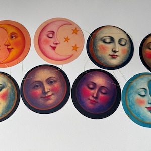 Op de afbeelding: Een decoratieve slinger met acht ronde papieren uitsneden, elk met een unieke hemelse illustratie van een maangezicht met verschillende uitdrukkingen en kleuren, waaronder tinten oranje, roze, blauw en paars, aan elkaar geregen.