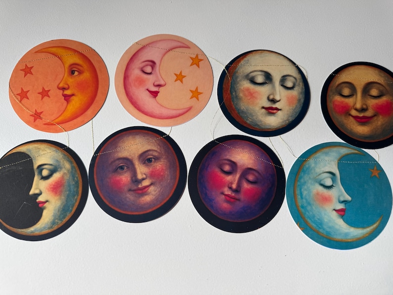 Op de afbeelding: Een decoratieve slinger met acht ronde papieren uitsneden, elk met een unieke hemelse illustratie van een maangezicht met verschillende uitdrukkingen en kleuren, waaronder tinten oranje, roze, blauw en paars, aan elkaar geregen.