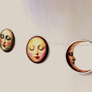 K&ouml;nnte beinhalten: Eine Girlande mit Mondphasen-Dekorationen. Jede runde Dekoration zeigt ein menschliches Gesicht, von Vollmond bis Sichel. Die Gesichter haben rosige Wangen und geschlossene Augen, im Vintage-Stil.