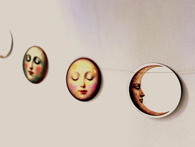 K&ouml;nnte beinhalten: Eine Girlande mit Mondphasen-Dekorationen. Jede runde Dekoration zeigt ein menschliches Gesicht, von Vollmond bis Sichel. Die Gesichter haben rosige Wangen und geschlossene Augen, im Vintage-Stil.