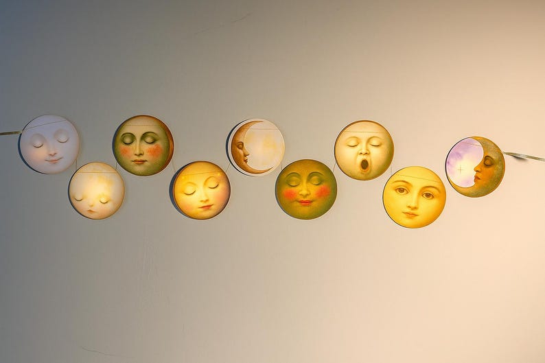 K&ouml;nnte beinhalten: Eine Lichterkette mit dekorativen mondf&ouml;rmigen Lichtern mit bemalten Gesichtern. Die Monde variieren in der Farbe, von blassgelb bis goldfarben, und weisen unterschiedliche Ausdr&uuml;cke auf. Die Lichter sind auf einem d&uuml;nnen Draht aufgereiht und erzeugen eine skurrile Himmelsdarstellung.