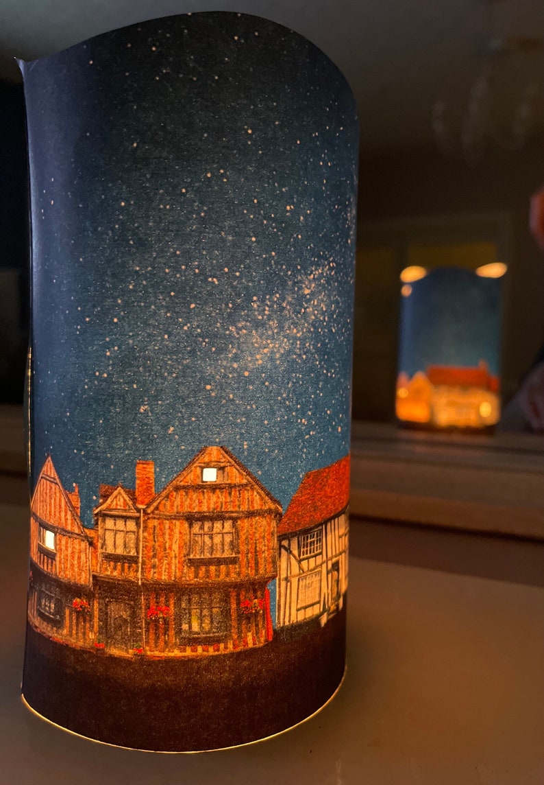 May include: A cylindrical lampshade with a starry night sky design. The lampshade features a row of three illuminated houses with a dark brown roof and white windows. The houses are set against a dark blue background with white stars.