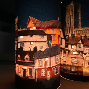 May include: Two cylindrical candle holders with a nighttime cityscape design. The design features a variety of buildings with red brick and white trim. The buildings are lit up with warm yellow lights.