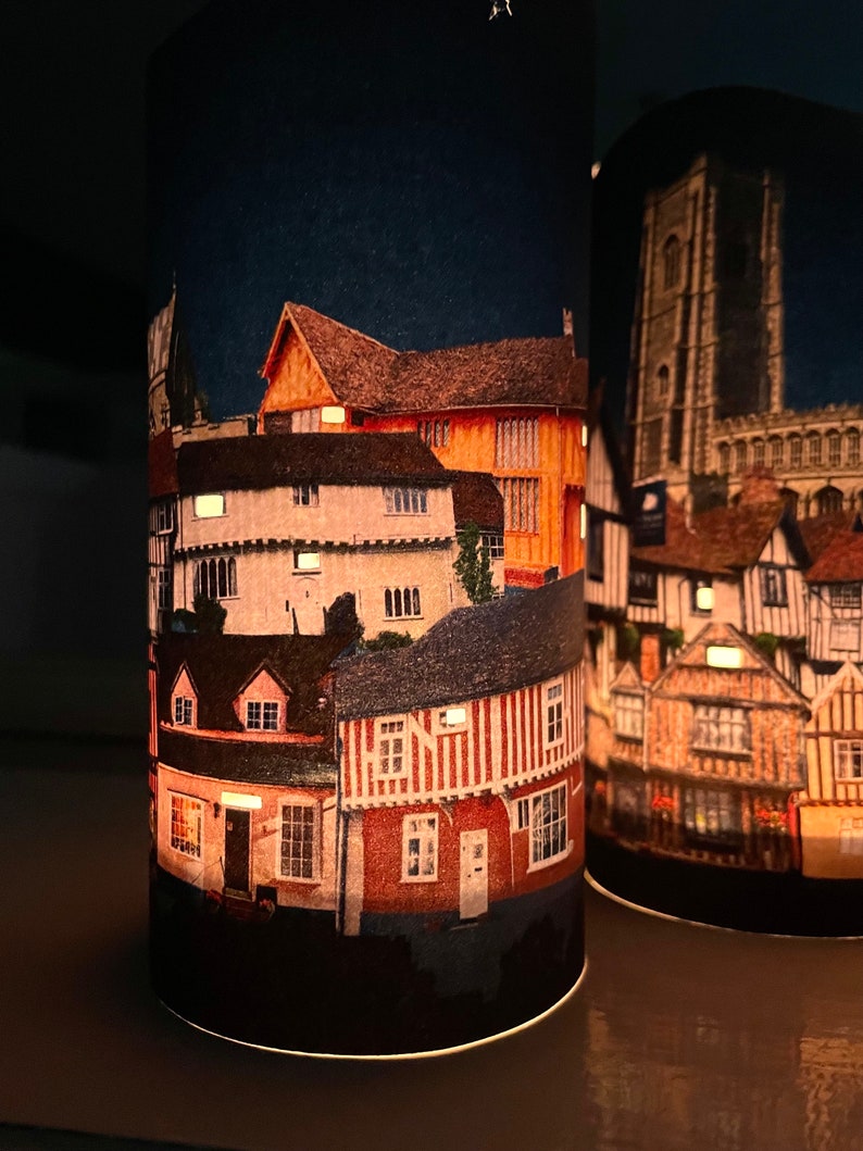 May include: Two cylindrical candle holders with a nighttime cityscape design. The design features a variety of buildings with red brick and white trim. The buildings are lit up with warm yellow lights.