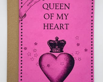 May include: A bright pink rectangular greeting card with a dark border of dots. The text reads "QUEEN OF MY HEART" with a banner above saying "ALWAYS". A large dark heart topped with a crown and surrounded by small stars is centred below.