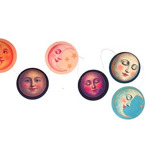Op de afbeelding: Een decoratieve slinger bestaande uit zeven ronde papieren uitsneden opgehangen aan een dun touwtje. Elke uitsnede toont een unieke illustratie van een maan met een menselijk gezicht, uitgevoerd in kleuren zoals oranje, roze, blauw en donkerblauw.