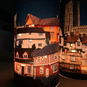 May include: Two cylindrical candle holders with a night scene of a town with buildings and a church tower. The candle holders are lit and the scene is illuminated.
