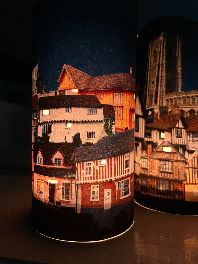 May include: Two cylindrical candle holders with a night scene of a town with buildings and a church tower. The candle holders are lit and the scene is illuminated.