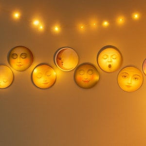 K&ouml;nnte beinhalten: Eine Sammlung von neun kreisf&ouml;rmigen Wanddekorationen, die Mondphasen darstellen, jede mit einem stilisierten Gesicht. Die Gesichter sind in warmen Farben bemalt und beleuchtet, mit einer Lichterkette dar&uuml;ber.