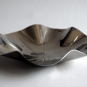 Puede incluir: Un cuenco de metal plateado con un borde ondulado y rizado. El cuenco est&aacute; hecho de metal pulido y refleja la luz.