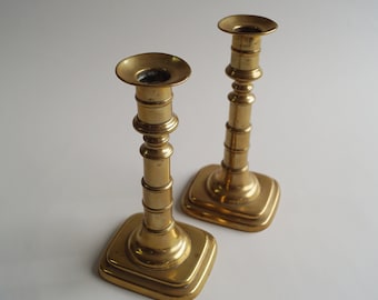 May include: Two gold metal candle holders with a simple design. The candle holders have a square base and a cylindrical stem with a flared top.