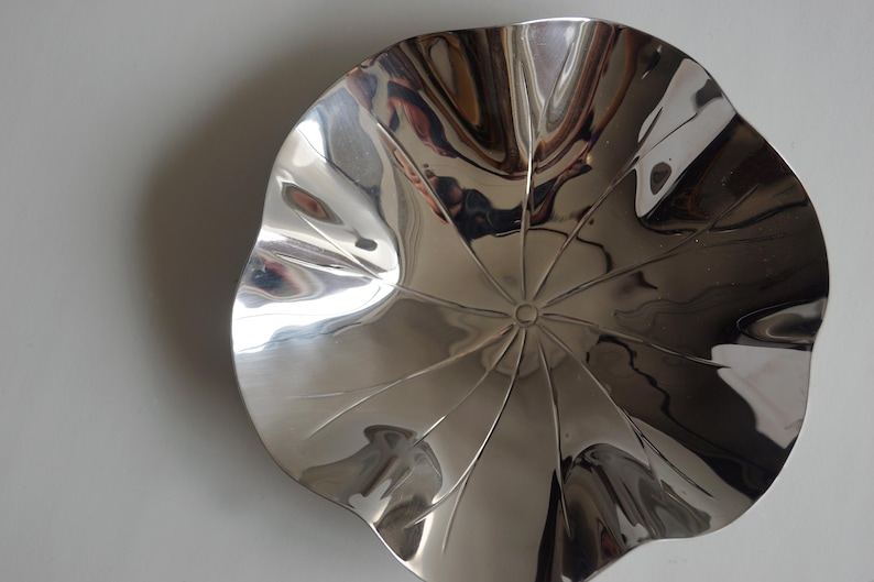 Puede incluir: Un cuenco decorativo de plata brillante con forma de flor y un centro texturizado. El cuenco est&aacute; hecho de metal y tiene una superficie lisa y reflectante.