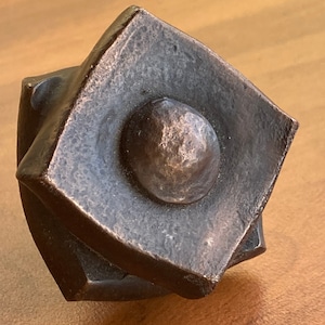 May include: A bronze-colored, geometrically shaped door knob with a round, raised center. The knob has a textured surface and a slightly worn appearance.