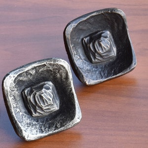 May include: Two square-shaped metal drawer pulls with a hammered finish. The pulls have a raised, textured design in the center.