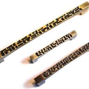 May include: Three bronze-toned cabinet pulls with a geometric, textured design. The pulls vary in size, with a long pull and two shorter ones. The hardware is set against a white background, highlighting the intricate details and finish.