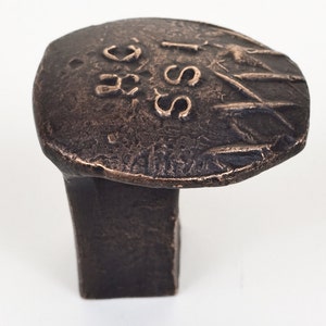 May include: A bronze-colored cabinet knob with a rectangular base and a rounded, textured top. The top features embossed text and markings. The hardware is designed for furniture and home decor.