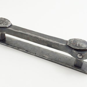 May include: Hand-forged iron door pull with a textured, dark gray finish. The handle features decorative, riveted end pieces and a rectangular bar. The mounting plate has pre-drilled holes for easy installation.
