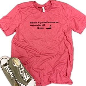 May include: A pink heather t-shirt with the text "Believe in yourself even when no one else will. -Nessie" and a black silhouette of the Loch Ness Monster.