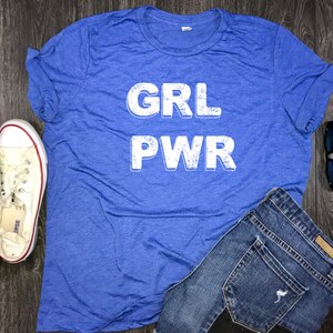 May include: A blue t-shirt with the words "GRL PWR" printed in white on the front. The shirt is laid flat on a wooden surface with a pair of white sneakers, a pair of blue jeans, and a pair of sunglasses.