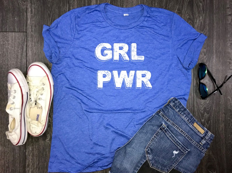 May include: A blue t-shirt with the words "GRL PWR" printed in white on the front. The shirt is laid flat on a wooden surface with a pair of white sneakers, a pair of blue jeans, and a pair of sunglasses.