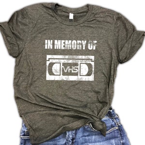 May include: A dark gray t-shirt with the text "IN MEMORY OF" and a vintage-style illustration of a VHS cassette tape.