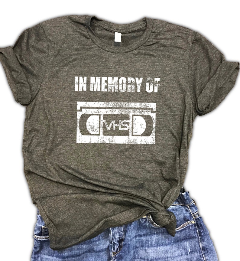 May include: A dark gray t-shirt with the text "IN MEMORY OF" and a vintage-style illustration of a VHS cassette tape.