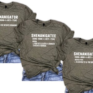 Puede incluir: Tres camisetas grises con texto blanco que definen las palabras "Shenanigator", "Shenanigatee" y "Shenanigated". Cada camiseta tiene una definici&oacute;n y una gu&iacute;a de pronunciaci&oacute;n. Las camisetas se muestran sobre un fondo de mezclilla.