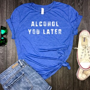 May include: A blue t-shirt with the text "Alcohol You Later" in white lettering. The shirt is tied in a knot at the front and is paired with denim shorts and white sneakers.