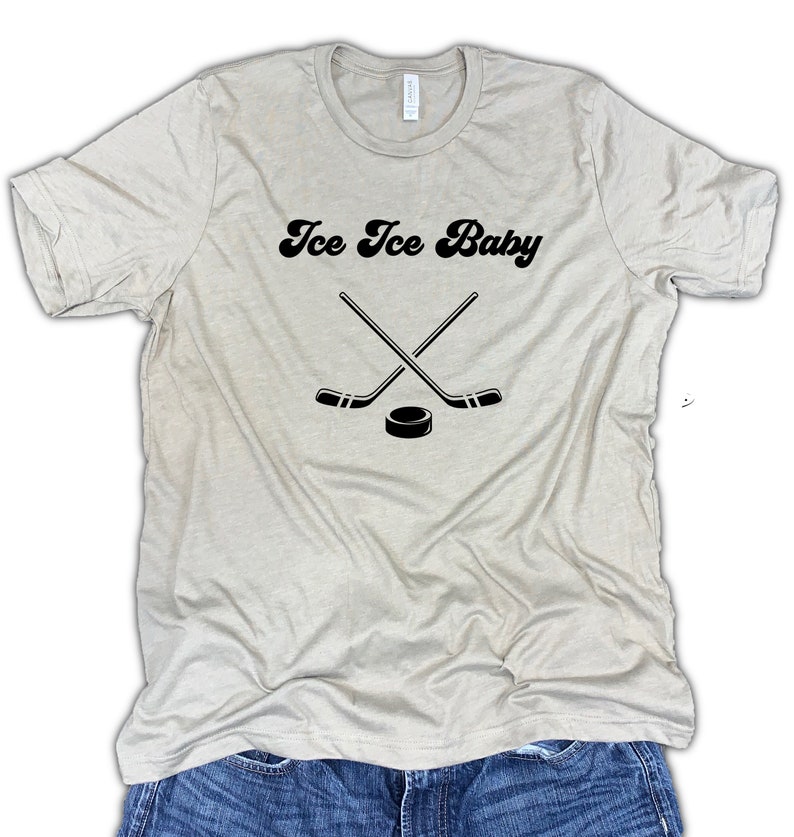 May include: A light beige t-shirt with the text "Ice Ice Baby" in a retro font. Two crossed hockey sticks and a puck are below the text.