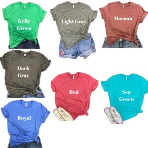 Pode incluir: Uma vista de cima de dez camisetas de diferentes cores, incluindo verde Kelly, cinza claro, vinho, cinza escuro, vermelho, verde mar e azul royal. Cada camiseta tem uma cor diferente e uma etiqueta de texto com o nome da cor.