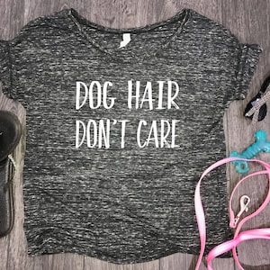 Puede incluir: Camiseta gris jaspeada de cuello en V con el texto "Dog Hair Don't Care" en blanco. La camiseta est&aacute; sobre una superficie de madera con una correa rosa, un juguete para perros azul y gafas de sol.
