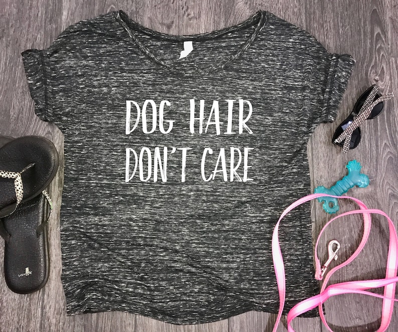 Puede incluir: Camiseta gris jaspeada de cuello en V con el texto "Dog Hair Don't Care" en blanco. La camiseta est&aacute; sobre una superficie de madera con una correa rosa, un juguete para perros azul y gafas de sol.
