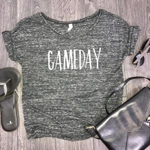 May include: A black and grey heather t-shirt with a white v-neck and the word "GAMEDAY" printed in white on the front.